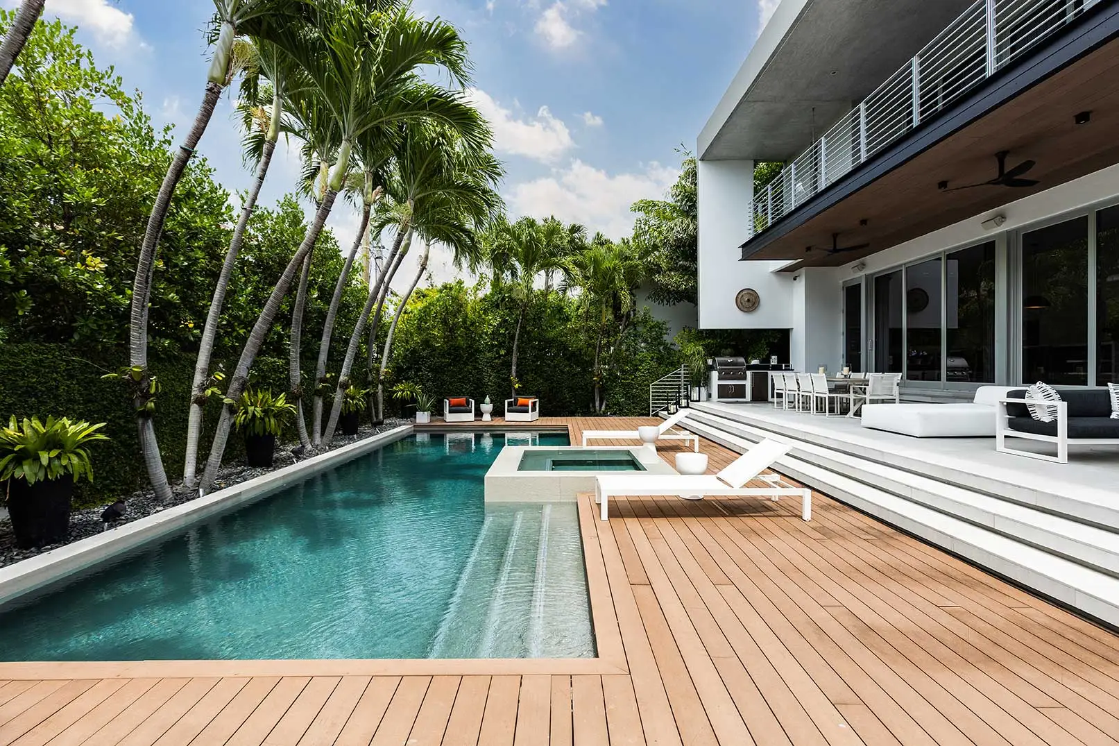 Luxurious backyard with a sleek pool surrounded by lush greenery and tall palm trees. Modern patio with white lounge chairs conveys relaxation.