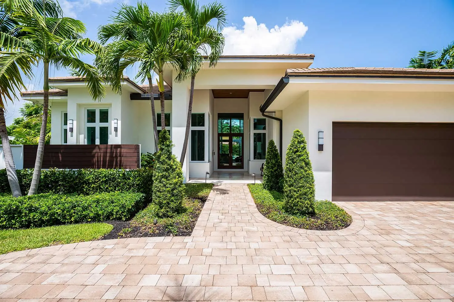Modern house exterior with white walls, large windows, and a brown garage door. Palm trees and green shrubs line the brick pathway, under a bright blue sky.